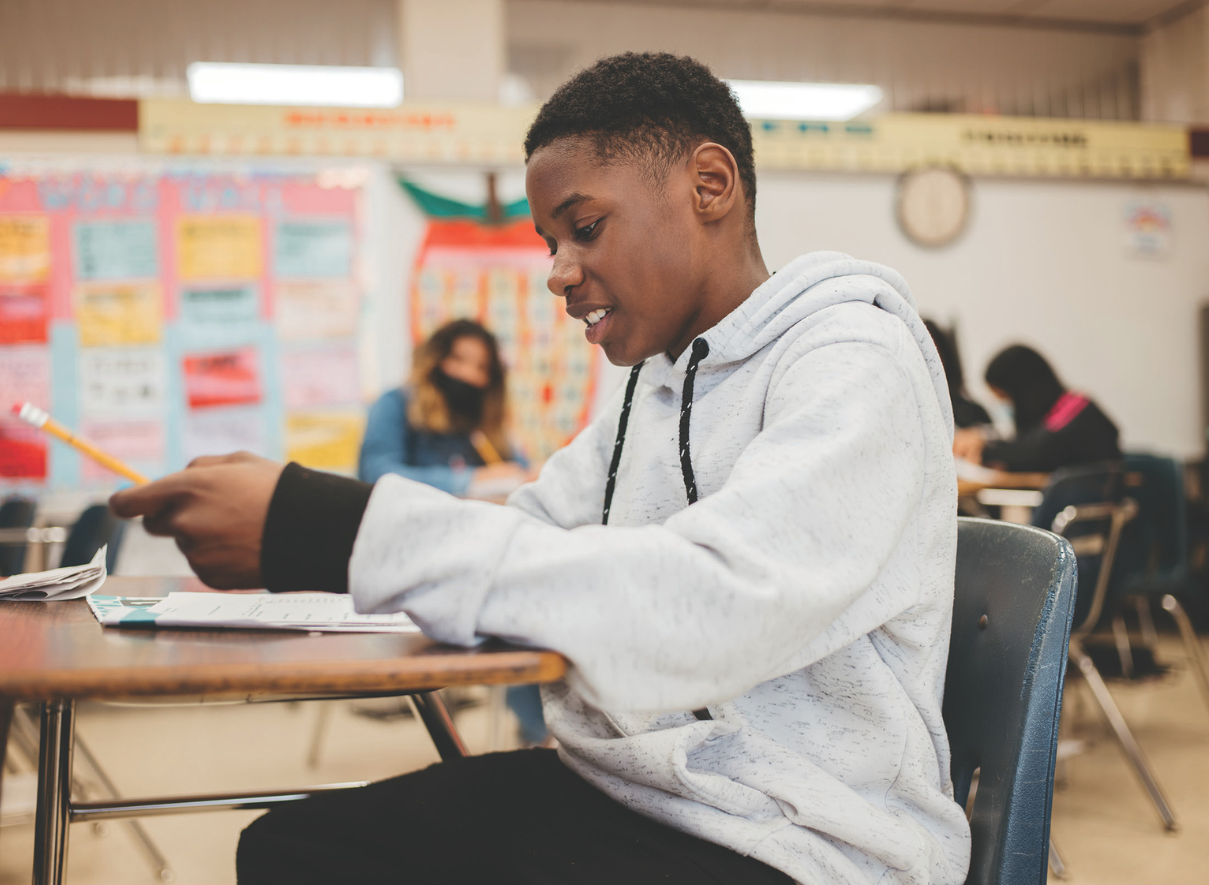 A young student wearing a light gray hoodie sits at a desk in a classroom, focused on reading or writing in papers in front of him. 